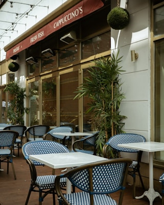 An outdoor café area with blue and white checkered chairs and white square tables. The café has a red awning with signs displaying 'Pastas', 'Fresh Fish', and 'Cappuccino's'. Green plants are placed along the walls and string lights are hung above.