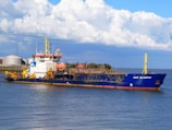 A large vessel named UKD Bluefin is sailing in a body of water. The ship is painted blue with a white and red superstructure and features yellow cranes and equipment. In the background, there are industrial buildings and storage tanks near the shoreline, and the sky is filled with large, fluffy clouds.