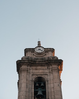 A stone clock tower with an analog clock face showing approximately 9:03. The tower features architectural details and two large bells visible through a window opening. The sky is clear and pale, suggesting early morning or late afternoon lighting.
