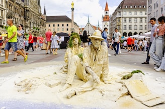 A street scene with two performers sitting on the ground covered in sand, creating the illusion of being made of stone. They are adorned with hats and greenery, resembling sculptures. The busy square is filled with people walking, surrounded by historic buildings with intricate facades and spires. The atmosphere is lively with onlookers passing by.