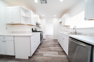 A modern, bright kitchen with white cabinetry and marble countertops. The room features stainless steel appliances, including a dishwasher and a stove, and has a wooden floor. Recessed lighting is installed in the ceiling, and natural light comes in through a large window with blinds.