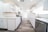 A modern, bright kitchen with white cabinetry and marble countertops. The room features stainless steel appliances, including a dishwasher and a stove, and has a wooden floor. Recessed lighting is installed in the ceiling, and natural light comes in through a large window with blinds.