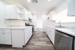 A modern, bright kitchen with white cabinetry and marble countertops. The room features stainless steel appliances, including a dishwasher and a stove, and has a wooden floor. Recessed lighting is installed in the ceiling, and natural light comes in through a large window with blinds.