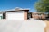 A single-story house with a beige stucco exterior and a white garage door. The driveway is spacious with a smooth concrete surface, and there is a tree with green leaves and small yellow flowers to the right. The roof features a classic gable design.