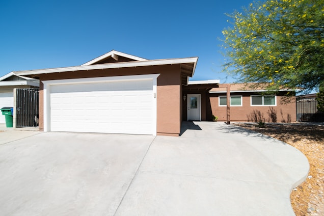 A single-story house with a beige stucco exterior and a white garage door. The driveway is spacious with a smooth concrete surface, and there is a tree with green leaves and small yellow flowers to the right. The roof features a classic gable design.