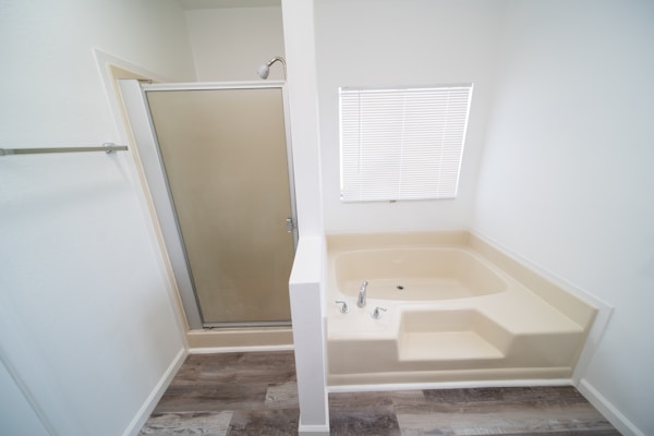 A bathroom featuring a beige corner bathtub with a step and a chrome faucet. To the left, there is a walk-in shower with a frosted glass door. Above the bathtub, a window with closed blinds allows diffused light into the space. The floor is covered with dark wood-patterned laminate.