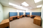 A spacious office room with several wooden desks arranged along the walls. Each desk features a blue partition for privacy, typical of a modern office setup. The room is well-lit with ceiling lights and has a window letting in natural light along one wall.