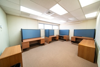 A spacious office room with several wooden desks arranged along the walls. Each desk features a blue partition for privacy, typical of a modern office setup. The room is well-lit with ceiling lights and has a window letting in natural light along one wall.