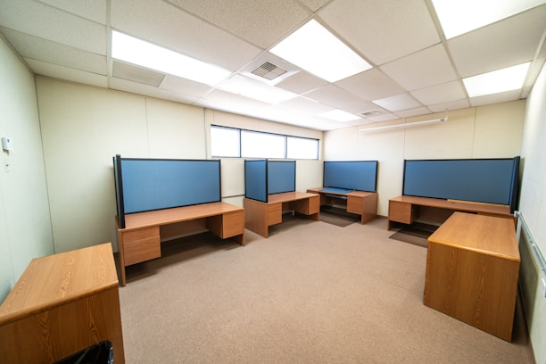 A spacious office room with several wooden desks arranged along the walls. Each desk features a blue partition for privacy, typical of a modern office setup. The room is well-lit with ceiling lights and has a window letting in natural light along one wall.
