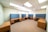 A spacious office room with several wooden desks arranged along the walls. Each desk features a blue partition for privacy, typical of a modern office setup. The room is well-lit with ceiling lights and has a window letting in natural light along one wall.