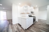 A modern kitchen featuring white cabinetry and a stainless steel stove. The countertops are light-colored, matching the overall bright and clean aesthetic. The flooring is a light wood laminate, which extends into the adjacent dining area. Vertical blinds cover the sliding glass door, allowing natural light to enter.