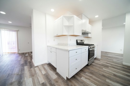 A modern kitchen featuring white cabinetry and a stainless steel stove. The countertops are light-colored, matching the overall bright and clean aesthetic. The flooring is a light wood laminate, which extends into the adjacent dining area. Vertical blinds cover the sliding glass door, allowing natural light to enter.