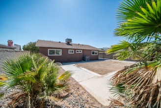 A single-story house with a brown exterior, surrounded by a minimal desert-style yard featuring palm trees and sparse vegetation. The yard has a patch of bare earth and a paved pathway leading up to the building. The sky is clear and blue, suggesting a sunny day.