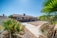 A single-story house with a brown exterior, surrounded by a minimal desert-style yard featuring palm trees and sparse vegetation. The yard has a patch of bare earth and a paved pathway leading up to the building. The sky is clear and blue, suggesting a sunny day.