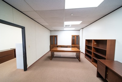 An empty office space with beige carpet and white walls. It features a large wooden desk at the center, flanked by wooden bookshelves and a cabinet with glass doors in the back. The room is well-lit by fluorescent ceiling lights.