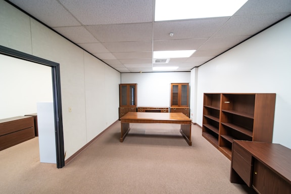 An empty office space with beige carpet and white walls. It features a large wooden desk at the center, flanked by wooden bookshelves and a cabinet with glass doors in the back. The room is well-lit by fluorescent ceiling lights.