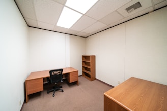 A small office room with beige walls and a brown carpet. The room features a wooden desk with a black swivel chair, a wooden bookshelf against the wall, and a wooden cabinet in the foreground. The ceiling has recessed fluorescent lighting.