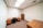 A small office room with beige walls and a brown carpet. The room features a wooden desk with a black swivel chair, a wooden bookshelf against the wall, and a wooden cabinet in the foreground. The ceiling has recessed fluorescent lighting.