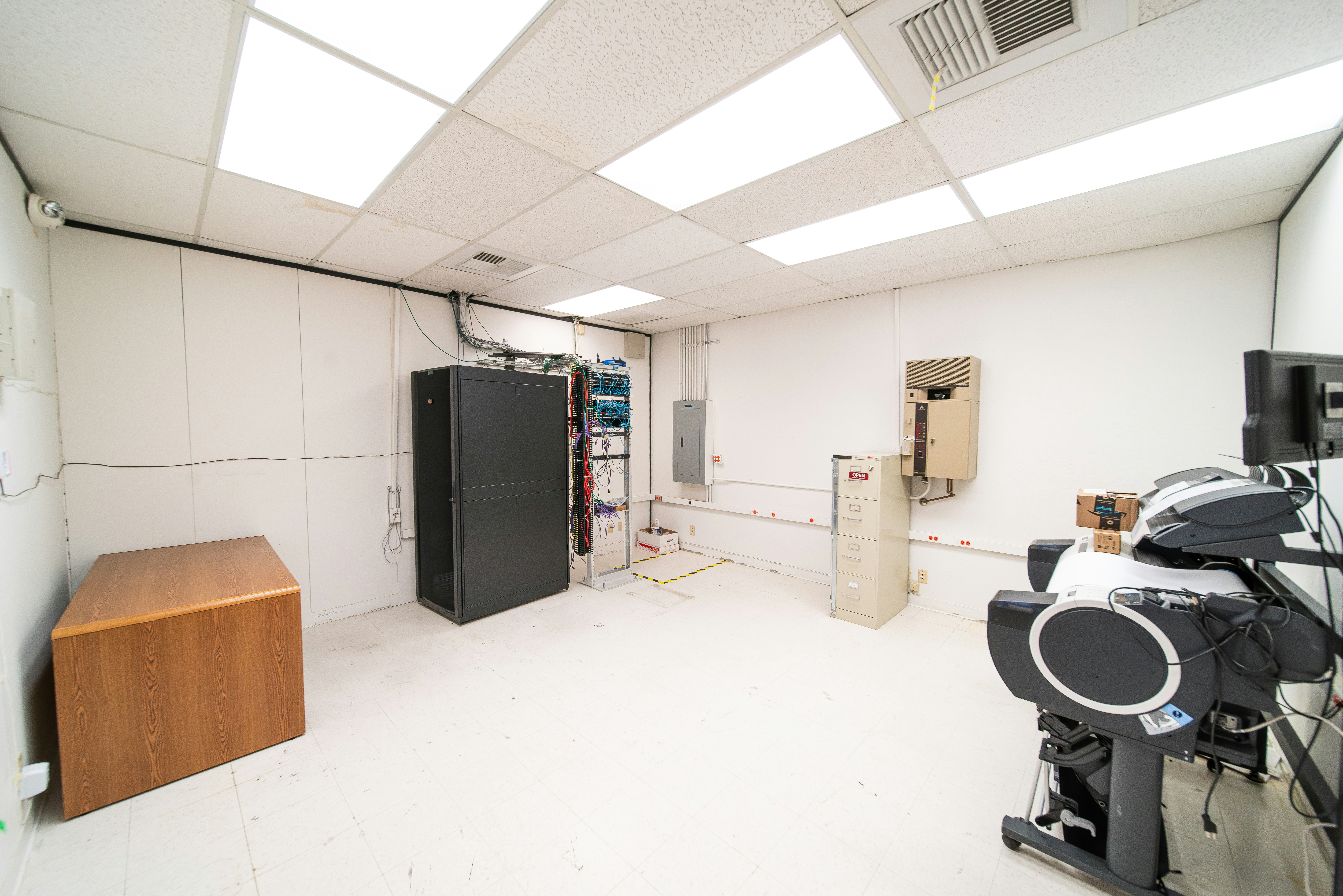 A room with a tiled ceiling and fluorescent lighting contains various electronic and office equipment. On the left, there's a wooden desk with a black server cabinet next to it. In the center of the room, a rack of network cables is visible, organized into a larger unit. To the right, a filing cabinet is mounted on the wall, with a printer/copier machine near the foreground. The walls are plain and the floor is covered in white tiles.