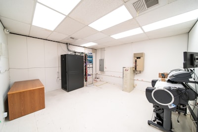 A room with a tiled ceiling and fluorescent lighting contains various electronic and office equipment. On the left, there's a wooden desk with a black server cabinet next to it. In the center of the room, a rack of network cables is visible, organized into a larger unit. To the right, a filing cabinet is mounted on the wall, with a printer/copier machine near the foreground. The walls are plain and the floor is covered in white tiles.