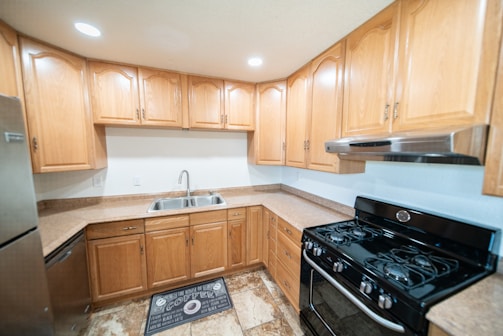 A modern kitchen features natural wood cabinets with a U-shaped configuration. The countertops are brown and complement the beige tiled floor. A stainless steel double sink with a chrome faucet is centrally located under the cabinets, while a black gas stove with an overhead range hood is on the right. Recessed ceiling lights brighten the space, and a dark kitchen mat is placed on the floor.
