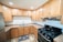 A modern kitchen features natural wood cabinets with a U-shaped configuration. The countertops are brown and complement the beige tiled floor. A stainless steel double sink with a chrome faucet is centrally located under the cabinets, while a black gas stove with an overhead range hood is on the right. Recessed ceiling lights brighten the space, and a dark kitchen mat is placed on the floor.