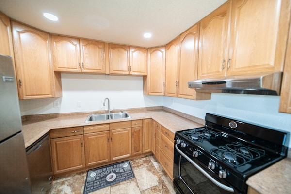 A modern kitchen features natural wood cabinets with a U-shaped configuration. The countertops are brown and complement the beige tiled floor. A stainless steel double sink with a chrome faucet is centrally located under the cabinets, while a black gas stove with an overhead range hood is on the right. Recessed ceiling lights brighten the space, and a dark kitchen mat is placed on the floor.