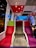 A modern train interior with vibrant multicolored seating. The seats are upholstered in red, green, gray, and purple fabrics with a dotted pattern. Overhead panels are in contrasting shades of red and gray. Soft lighting from ceiling fixtures adds a warm ambiance. Large windows are visible on the side, allowing a peek outside to a blue-lit exterior. The train is spacious with clean, sleek surfaces.