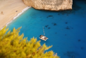 A serene cove featuring a small beach with golden sand and clear blue water. A solitary sailboat floats on the water near the coastline, and there are some scattered, dark shadows visible underwater. The scene is framed by a lush, green bush in the foreground and rugged cliffs in the background.