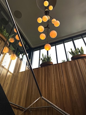 A modern interior space featuring a staircase with metal railings and wooden paneling. Above, several spherical pendant lights emit a warm glow. Large windows provide ample natural light, and potted plants add a touch of greenery.