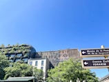 An urban scene with a building covered in lush greenery. The clear blue sky provides a backdrop, while signage directs to cultural and postal museums. The architecture combines natural elements with urban structures, showcasing a mix of modern and traditional styles.