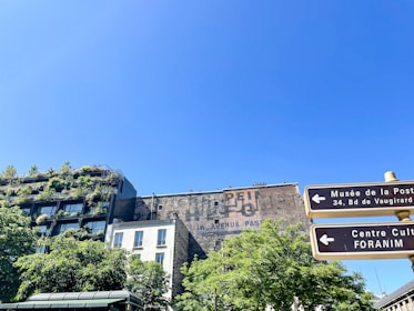 An urban scene with a building covered in lush greenery. The clear blue sky provides a backdrop, while signage directs to cultural and postal museums. The architecture combines natural elements with urban structures, showcasing a mix of modern and traditional styles.