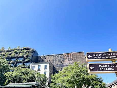 An urban scene with a building covered in lush greenery. The clear blue sky provides a backdrop, while signage directs to cultural and postal museums. The architecture combines natural elements with urban structures, showcasing a mix of modern and traditional styles.