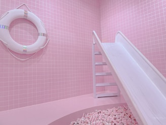 A pastel pink room features a life preserver mounted on a tiled wall and a white slide leading into a pit filled with pink and white marshmallows. The overall setting is whimsical and playful, with a monochromatic pink theme.