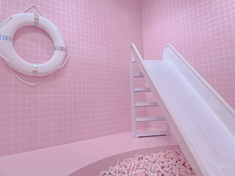 A pastel pink room features a life preserver mounted on a tiled wall and a white slide leading into a pit filled with pink and white marshmallows. The overall setting is whimsical and playful, with a monochromatic pink theme.
