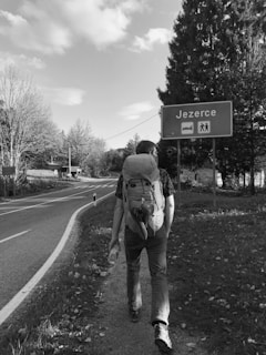 A person with a large backpack is walking along a road bordered by trees and grass. A sign indicates the direction to a place named 'Jezerce' which appears to offer accommodations and a hiking area. The scene is captured in black and white, adding a classic and nostalgic feel.