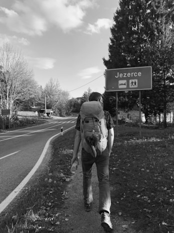 A person with a large backpack is walking along a road bordered by trees and grass. A sign indicates the direction to a place named 'Jezerce' which appears to offer accommodations and a hiking area. The scene is captured in black and white, adding a classic and nostalgic feel.