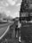 A person with a large backpack is walking along a road bordered by trees and grass. A sign indicates the direction to a place named 'Jezerce' which appears to offer accommodations and a hiking area. The scene is captured in black and white, adding a classic and nostalgic feel.