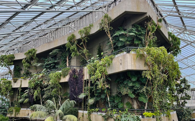 A concrete structure surrounded by lush green plants and trees, housed within a large greenhouse with a glass-paneled roof. The upper levels of the building have dense foliage, adding a vibrant, natural feel to the otherwise industrial environment.