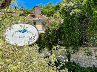 A rustic sign with the image of plums and the words 'La Prunière' hangs among vibrant foliage. Stone walls and a quaint house with a chimney can be seen partially obscured by lush greenery and blooming flowers, under a bright blue sky.