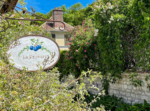A rustic sign with the image of plums and the words 'La Prunière' hangs among vibrant foliage. Stone walls and a quaint house with a chimney can be seen partially obscured by lush greenery and blooming flowers, under a bright blue sky.