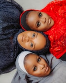 Three individuals are lying down with their heads close together, each wearing a different colored hijab. The hijabs are intricately styled with textures and patterns. The colors include dark blue, bright red, and light gray. The image captures their direct gaze, highlighting expressive makeup and elegant features.