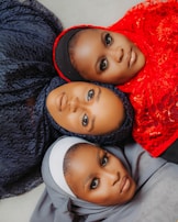 Three individuals are lying down with their heads close together, each wearing a different colored hijab. The hijabs are intricately styled with textures and patterns. The colors include dark blue, bright red, and light gray. The image captures their direct gaze, highlighting expressive makeup and elegant features.