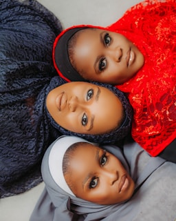 Three individuals are lying down with their heads close together, each wearing a different colored hijab. The hijabs are intricately styled with textures and patterns. The colors include dark blue, bright red, and light gray. The image captures their direct gaze, highlighting expressive makeup and elegant features.