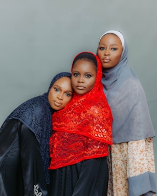 Three women are dressed in hijabs, standing closely together against a plain background. Each woman wears a different colored hijab: black, red, and light blue. They appear calm and composed, with the central figure wearing a red hijab standing slightly in front.