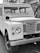 A vintage Land Rover with a classic design, featuring a boxy shape and large round headlights. The vehicle is parked with visible wear indicating its age. Its grille is prominently displaying the brand name. The setting includes a paved surface and a building in the background.