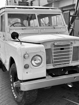 A vintage Land Rover with a classic design, featuring a boxy shape and large round headlights. The vehicle is parked with visible wear indicating its age. Its grille is prominently displaying the brand name. The setting includes a paved surface and a building in the background.