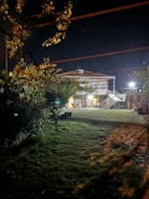 A nighttime scene featuring a two-story house with illuminated exterior. A grass lawn extends in the foreground, bordered by trees and shrubs. Outdoor lighting casts shadows on the ground, and a hammock is visible on the house's porch.