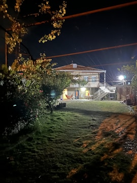 A nighttime scene featuring a two-story house with illuminated exterior. A grass lawn extends in the foreground, bordered by trees and shrubs. Outdoor lighting casts shadows on the ground, and a hammock is visible on the house's porch.