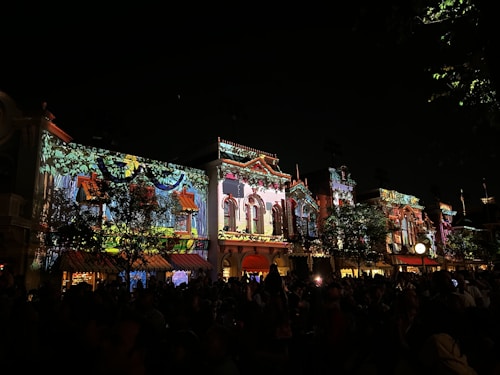 Vibrant light projections adorn the facades of traditional architectural buildings at night, creating a lively and colorful atmosphere. The crowd of people in the foreground adds to the festive energy.